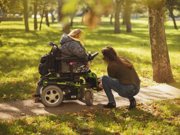 Osobná asistentka v parku počas jesene šnuruje topánku klientke na elektrickom vozíku.